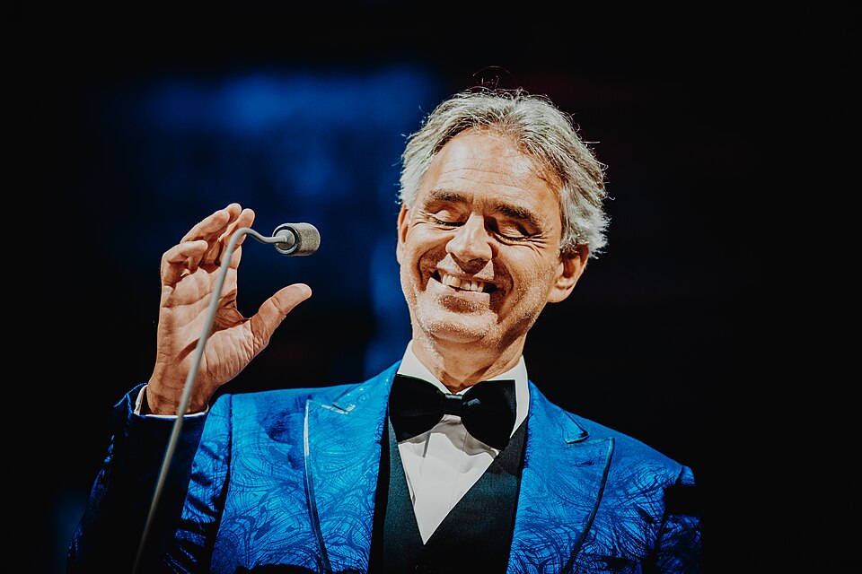 Andrea Bocelli in giacca blu e papillon nero, sorridente accanto al microfono con una mano alzata; fotografia a colori durante un concerto
