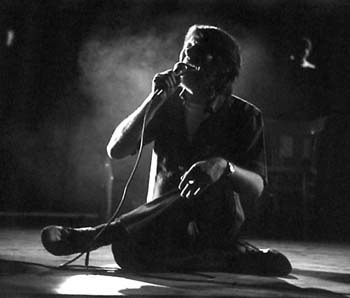 Fabrizio De André seduto sul palco a gambe incrociate, con un microfono in mano, in una fotografia in bianco e nero