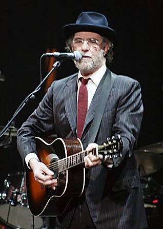 Francesco De Gregori al microfono con chitarra acustica, cappello scuro, occhiali e abito gessato