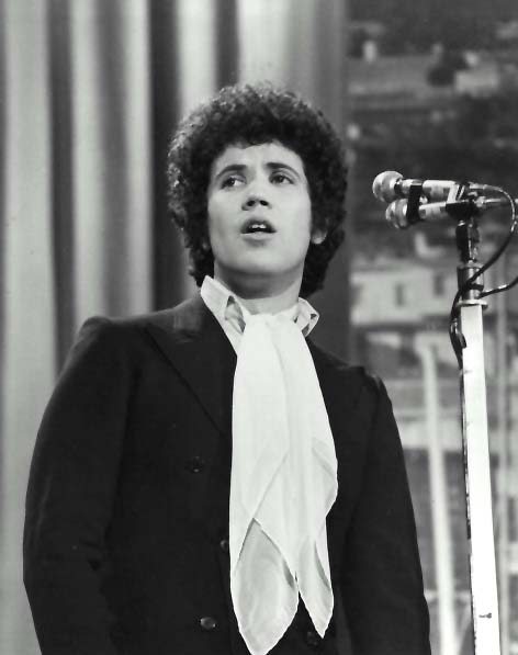 Lucio Battisti sul palco di Sanremo nel 1969, in giacca scura e foulard chiaro davanti ai microfoni. Fotografia in bianco e nero.