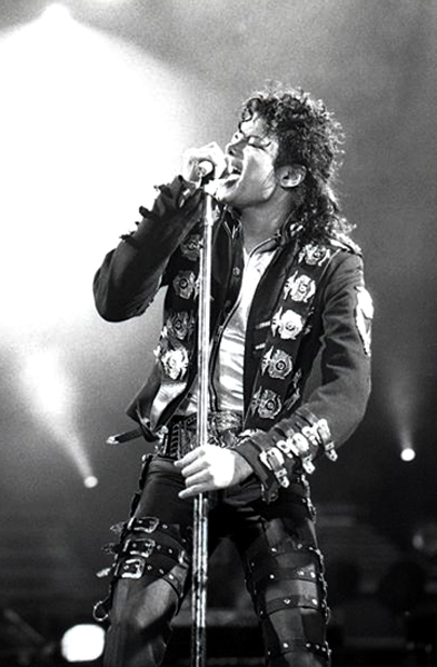 Michael Jackson canta al microfono sul palco, indossando una giacca scura con decorazioni chiare