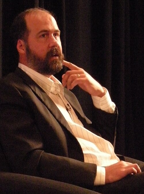 Il solo Krist Novoselic nel 2008, con barba, giacca scura e camicia chiara a righe, seduto con una mano vicino al viso; fotografia a colori