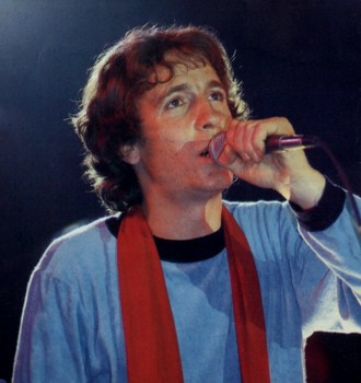Rino Gaetano con capelli mossi e una sciarpa rossa, mentre canta in un microfono tenuto nella mano sinistra; fotografia a colori su fondo scuro