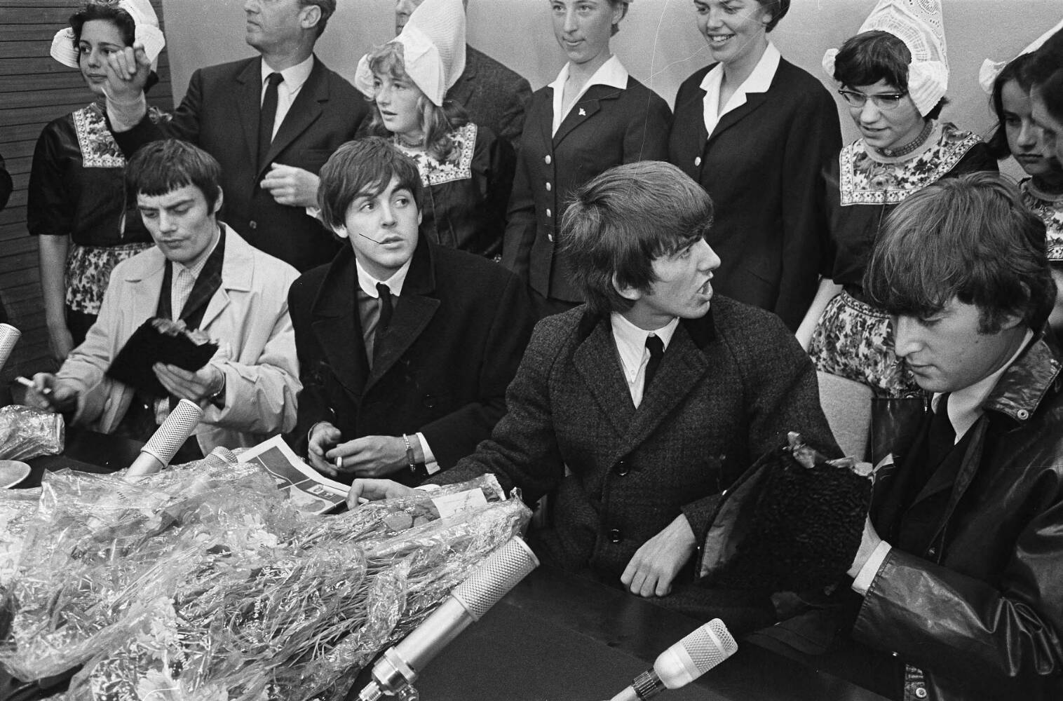 Jimmy Nicol, Paul McCartney, George Harrison e John Lennon seduti davanti ai microfoni a Schiphol nel 1964; Ringo Starr non è raffigurato. Fotografia in bianco e nero