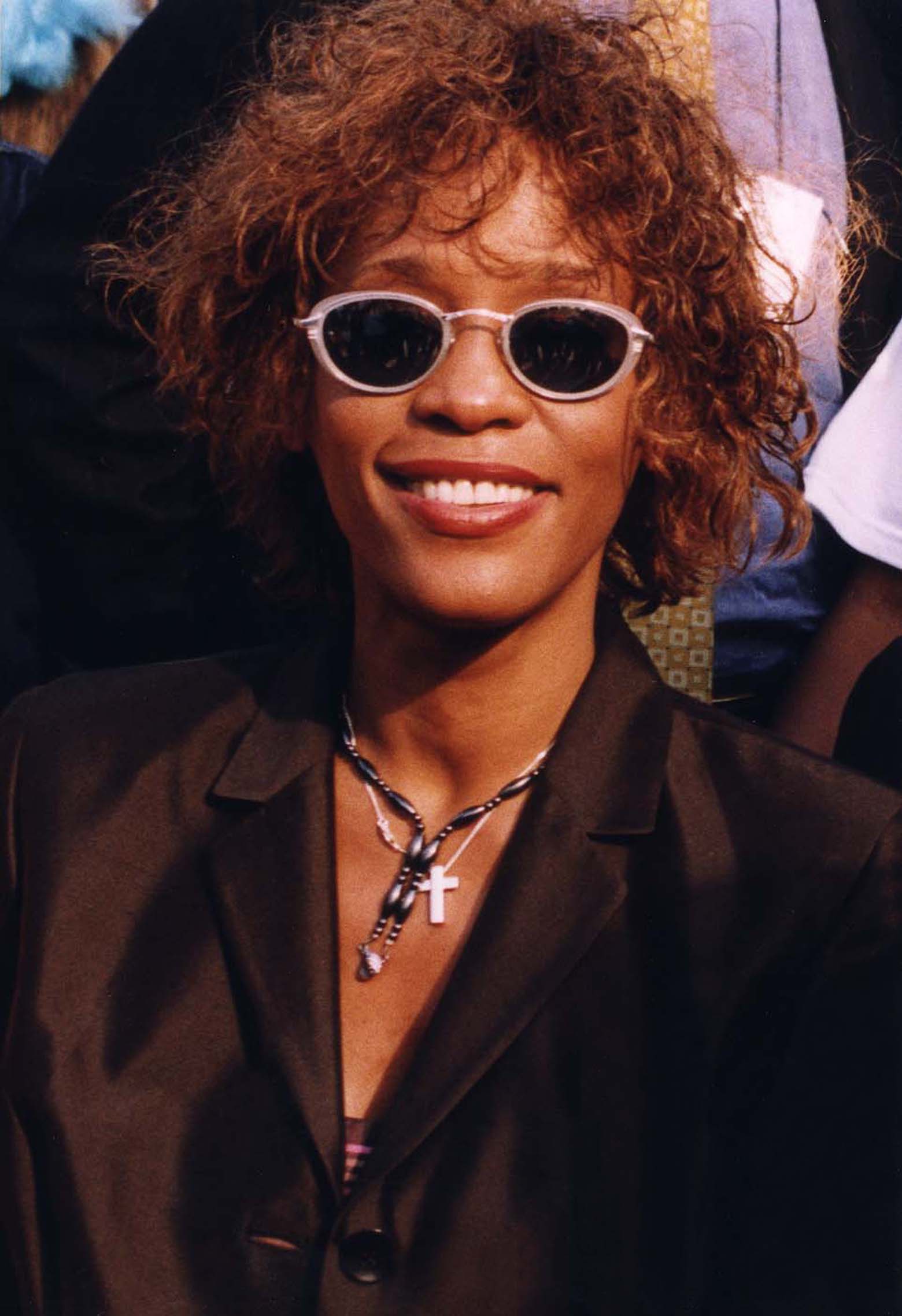 Whitney Houston sorride con occhiali ovali scuri e una giacca nera